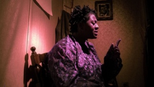 An African American woman in a purple shirt sits in a chair in a dark room, speaking to the audience.