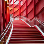 The red metal staircase of the Reginald F. Lewis Museum in Baltimore climbing between industrial-feeling red metal walls. Clear glass walls and silver metal rails line the staircase.