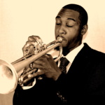 A young African-American man in a black suit, white shirt, and black tie with close-cropped hair closes his eyes while playing a trumpet. The sepia-colored black & white photo shows only his head and torso to his elbows, and has a very 1950s look and feel.