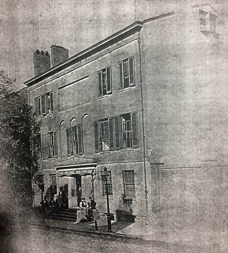 A very old photo of the Peale building, showing a three-story brick structure in people on the front porch. 