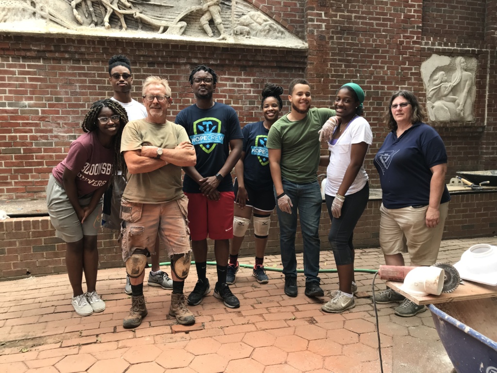 A group of eight young adults and a team leader pose for a photo by the brick wall in the Peale's garden. 