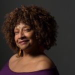 An African American woman, wearing dangling earrings and a purple off-the-shoulder top, smiles at the camera.