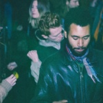 A vintage image of a man, wearing a black leather jacket, at a nightclub as people dance around him. 