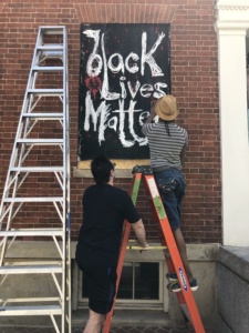 A white man holds a ladder for a black man who is installing a large "Black Lives Matter" painting on a boarded up window.