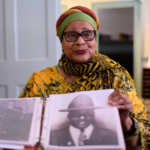 Mama Linda Goss wears a colorful turbin and holds a photo of a man in a fedora hat.