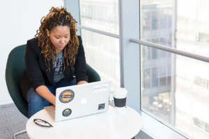 A woman with long braids and a blue blazer sits at a laptop computer in an office by a window.