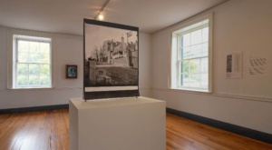 Gallery view of a room with light-colored hardwood floors and a black-and-white photograph on a pedestal in the middle.
