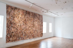 A white, contemporary gallery space with a large earth-toned mosiac on the wall, showing people's faces and bodies.