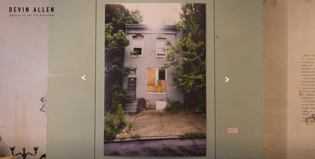 A view of a photograph in Devin Allen's Spaces of the Un-Entitled exhibition, showing a blue row house with boarded-up windows.