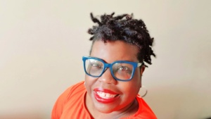 Daisy Brown wears a bright orange t-shirt and blue eyeglasses as she smiles up at the camera.