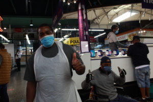 Two men wearing masks and aprons stand in the Market and give the thumbs up sign.