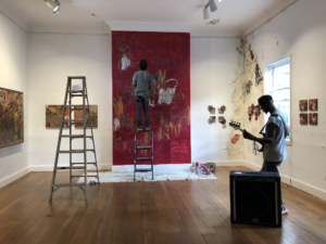 A man stands on a ladder and paints a large, red canvas as a man plays guitar in the background.