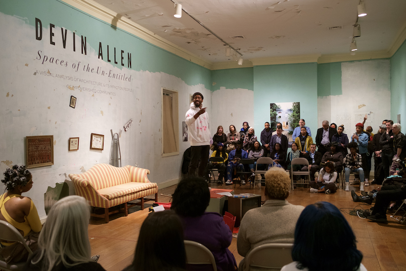 A man stands in front of a crowd of onlookers in a gallery space at a museum.
