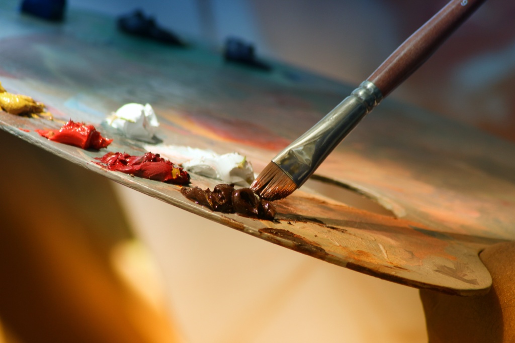 A close-up view of an artist's palette and a paint brush that it being dipped into a gob of dark brown paint.