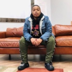 Chrys Seawood has short black hair and wears a jean jacket, green pants, and black Croc shoes. She sits on a brown leather couch.