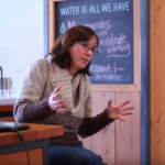 Heather wears a brown sweater, sits, on a bench, and has her hands out in gesture as she talks to a group.