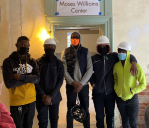 Five people stand in front of a sign that reads, Moses Williams Center. All of the people wear face masks and winter clothing. They stand in a historic space that is being renovated.