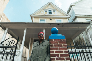 A woman in an army green jacket leans against a brick column, in front of a multi-story, cream-colored house.