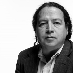 Paul Chaat Smith has wavy black hair which is pulled back. He wears a dark-colored blazer and light, buttoned down shirt.