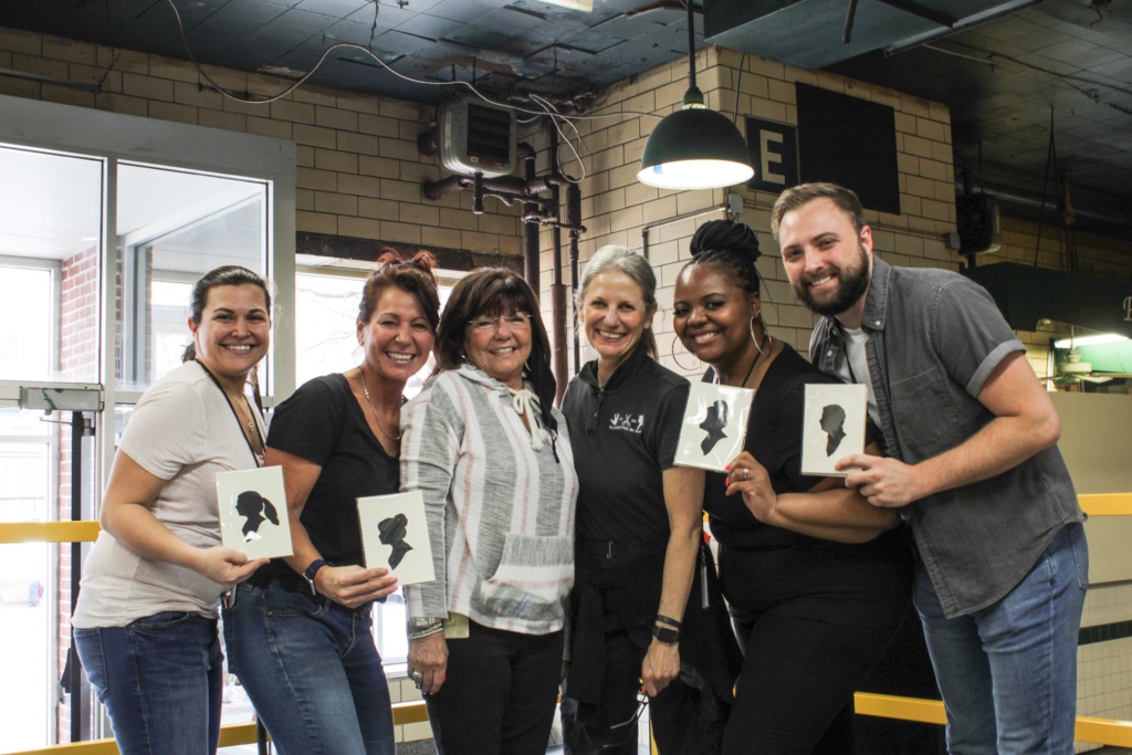 A group of individuals stands in a market space and holds up small black silhouette portraits of themselves.