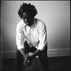 A man with a white collared shirt and eyeglasses sits on a stool with the backdrop of a blank wall.