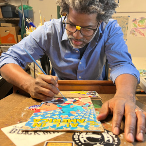 A man in a blue collared shirt and eyeglasses sits at a table and paints a comic book cover.