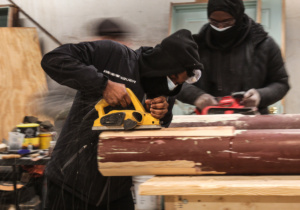 A man in a black sweatshirt uses a belt sander to sand down a large, wooden column.