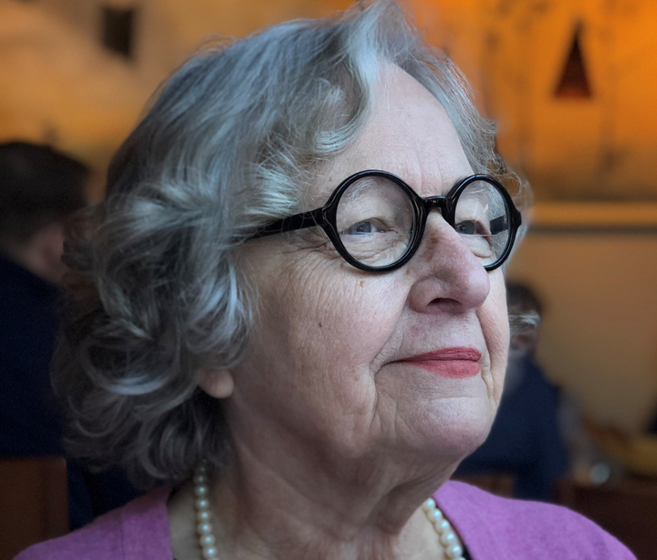 A woman with gray hair, round glasses, and a purple top stands in front of an orange-colored background.